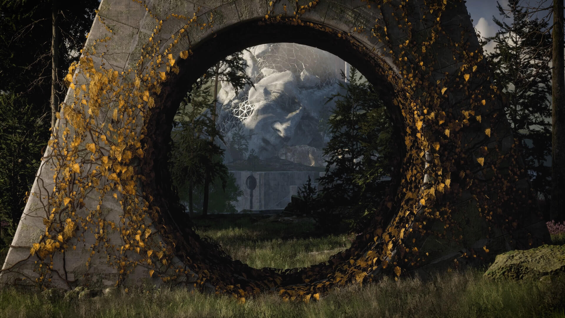 Paisagem de The Talos Principle 2 vista através de um grande arco de pedra envelhecida coberto por trepadeiras e folhas amarelas. No centro há uma floresta de coníferas e, ao fundo, uma enorme escultura de um rosto humano de pedra parcialmente coberta por estruturas geométricas brancas. O primeiro plano tem grama, terra e uma rocha com musgo, com luz solar atravessando partes da vegetação.